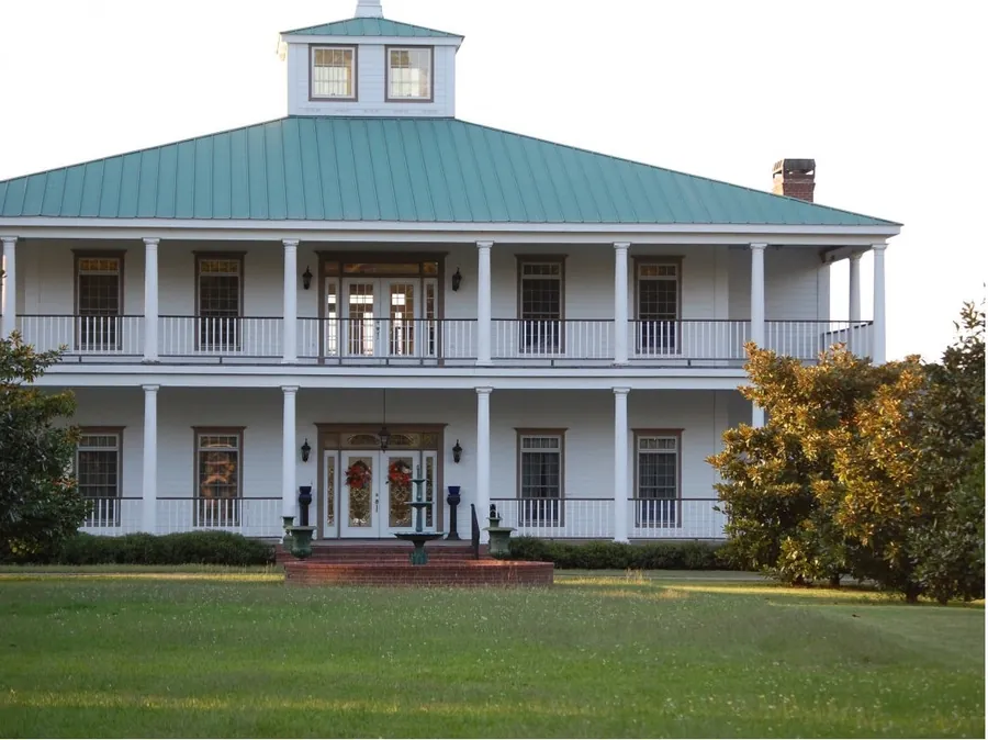 Two-story white home with full-width upper and lower galleries, a green metal roof and cupola, in warm evening light