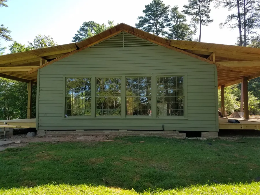 Green-sided addition with a wall of divided-light windows and a new wraparound porch roof under construction